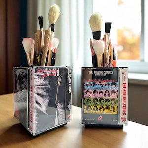 May include: Two square desk organizers constructed from clear plastic cassette tape cases. One features The Rolling Stones Tattoo You album art, the other Some Girls. Both are filled with various makeup brushes and wooden pencils.