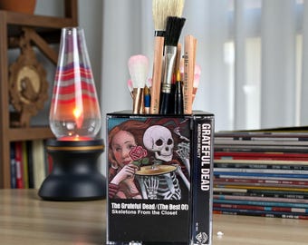 May include: A rectangular desk organizer constructed from a clear plastic cassette case featuring the Grateful Dead Skeletons From the Closet album cover art. The container is filled with various artist paintbrushes and wooden pencils.