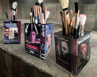 May include: Three square desk organizers constructed from vintage VHS cassette cases. Each container holds various makeup brushes and pens. The cases feature movie cover art for The Shining, A Nightmare on Elm Street, Halloween 5, and Halloween.