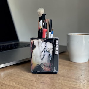 May include: A desk organizer made from a Talking Heads "Stop Making Sense" cassette tape. The organizer holds makeup brushes, pens, and a marker. The cassette tape has a black base and a clear plastic cover.