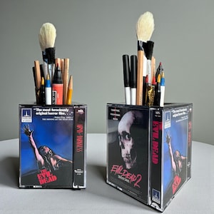 May include: Two clear acrylic pen holders made from repurposed VHS tapes. One features the movie "Army of Darkness" and the other "Evil Dead 2." Each holds various pens, pencils, and brushes, with a large makeup brush in each. The text "The Evil Dead" is visible.