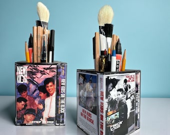May include: Two square pencil holders constructed from clear plastic cassette cases featuring New Kids on the Block album covers. Each holder is filled with various art supplies including wooden pencils, paint brushes, and markers.