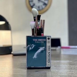 May include: A rectangular desk organizer constructed from a clear plastic cassette tape case featuring The Smiths album cover art for The Queen Is Dead. The holder contains various makeup brushes and pencils with wooden and black handles.