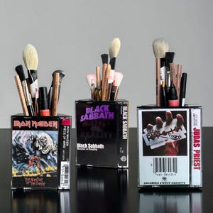May include: Three makeup brush holders made from repurposed cassette tape cases. The cases feature album art from Iron Maiden, Black Sabbath, and Judas Priest. Each holder is filled with various makeup brushes and tools, showcasing a unique and artistic storage solution.