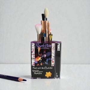 May include: A clear plastic cassette tape case repurposed as a makeup brush holder. The case features the album art for Prince and the Revolution's "Purple Rain" album. The holder contains various makeup brushes and a purple pencil.