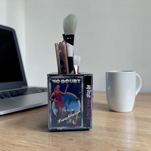 Peut inclure: Un récipient rectangulaire transparent avec les mots "No Doubt" et "Tragic Kingdom" imprimés, contenant des pinceaux de maquillage. Le récipient a un design vintage avec une femme en robe rouge. Une tasse blanche et un ordinateur portable sont en arrière-plan.