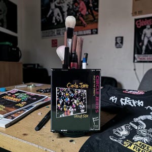 May include: A black cassette tape case repurposed as a makeup brush holder. The case features the band name "Circle Jerks" and album art. The holder contains various makeup brushes and applicators, with a black base.