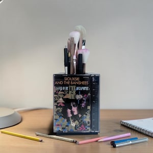 May include: A rectangular desk organizer repurposed from a clear plastic cassette tape case featuring the Siouxsie and the Banshees album cover for A Kiss in the Dreamhouse. The case is filled with various makeup brushes and writing pens.
