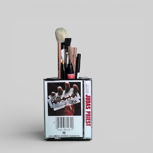 May include: A unique pencil holder made from a repurposed cassette tape. The tape features the album art for Judas Priest's "British Steel." The holder contains various makeup brushes and pencils, with the text "Judas Priest" visible on the side.