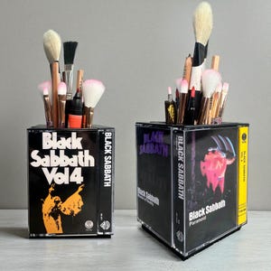 May include: Two desk organizers constructed from clear plastic cassette tape cases featuring Black Sabbath album artwork. The left case displays Vol 4, while the right case shows Paranoid and Master of Reality. Both are filled with various makeup brushes.