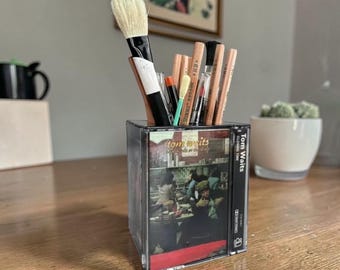 May include: A clear plastic cassette tape case repurposed as a pencil holder, containing various art brushes and pencils. The front of the case features the Tom Waits Closing Time album cover art, displaying a restaurant scene with red accents.