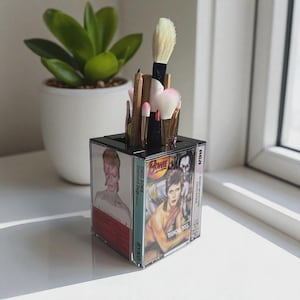 May include: A black and clear acrylic makeup brush holder featuring album art of David Bowie. The holder contains various makeup brushes and pencils. A small potted plant sits in the background.