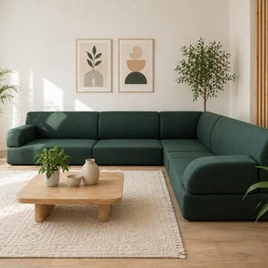 Op de afbeelding: Een moderne woonkamer met een grote, donkergroene hoekbank. De kamer bevat een lichtgekleurd tapijt, een houten salontafel en potplanten. Twee ingelijste kunstwerken hangen aan de witte muur en een raam is zichtbaar.