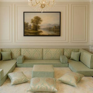 Op de afbeelding: Een ruime woonkamer met een grote, lichtgroene hoekbank en een bijpassende vierkante poef. Decoratieve kussens en een landschapsschilderij dragen bij aan de elegantie van de kamer. Een kroonluchter hangt aan het plafond.