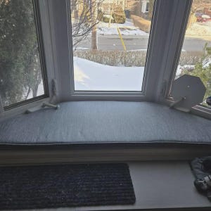 May include: A custom-fitted gray fabric window seat cushion with a textured weave, designed to sit flush within the curved frame of a bay window. The cushion features a clean, straight edge and a neutral gray tone that complements the window area.