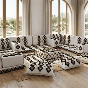 Puede incluir: Un juego de sala moderno con un diseño modular. Los sofás y cojines blancos presentan un patrón geométrico en negro y marrón. La habitación tiene grandes ventanales, pisos de madera y una paleta de colores neutros.