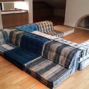 Può includere: Un divano componibile a forma di U composto da più cuscini con un motivo a righe blu e grigie. I cuscini sono realizzati in un tessuto morbido e confortevole.