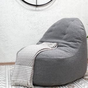 May include: A grey beanbag chair with a patterned throw blanket. The chair is in a room with a patterned rug, a small wooden side table, and potted plants. A decorative clock hangs on the wall.