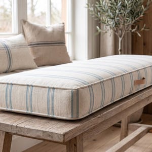 May include: A rectangular bench cushion featuring a cream-colored fabric with vertical light blue stripes. The cushion has piped edges and a small tan leather pull tab on the side, resting on a wooden bench with a textured, natural finish.