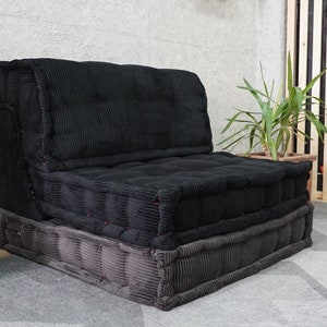 Puede incluir: Un sof&aacute; fut&oacute;n de terciopelo negro con una base gris. El sof&aacute; est&aacute; hecho de varios cojines y est&aacute; sentado sobre una alfombra gris.