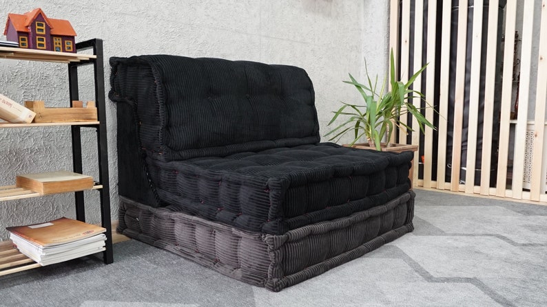 Puede incluir: Un sof&aacute; fut&oacute;n de terciopelo negro con una base gris. El sof&aacute; est&aacute; hecho de varios cojines y est&aacute; sentado sobre una alfombra gris.