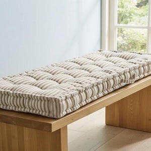 May include: A rectangular bench cushion with a striped pattern in beige and white, resting on a wooden bench. The cushion has a tufted design. The bench is made of light-colored wood. The scene is illuminated by natural light.