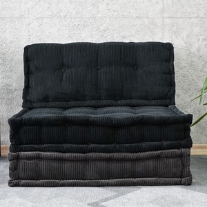 Puede incluir: Un coj&iacute;n de suelo de terciopelo de pana negro y gris con un respaldo capiton&eacute;. El coj&iacute;n est&aacute; sentado en una alfombra gris frente a una pared blanca.