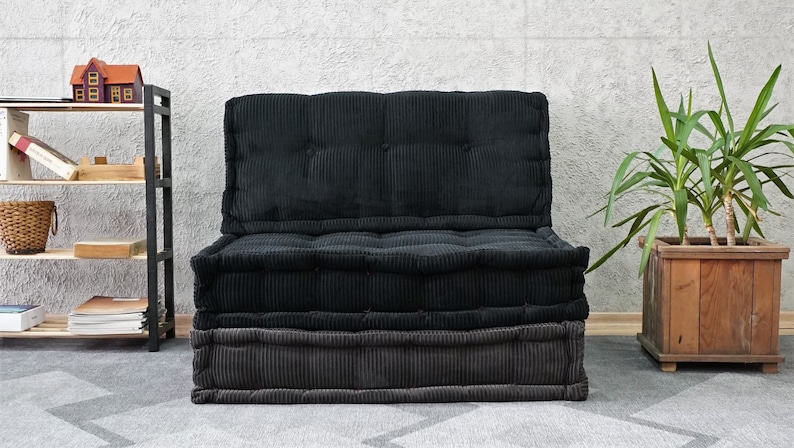 Puede incluir: Un coj&iacute;n de suelo de terciopelo de pana negro y gris con un respaldo capiton&eacute;. El coj&iacute;n est&aacute; sentado en una alfombra gris frente a una pared blanca.