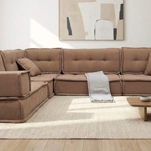 Könnte beinhalten: Ein modulares, braunes Ecksofa mit getufteten Kissen und Armlehnen. Das Sofa ist L-förmig angeordnet und steht auf einem hellen Teppich. Ein Couchtisch aus Holz und dekorative Gegenstände befinden sich vor dem Sofa.