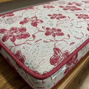 May include: A rectangular cushion with a floral pattern in shades of red and white. The cushion has a red border and is placed on a wooden bench. The floral design features large, stylized flowers and leaves.