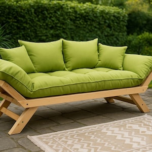 May include: A light green futon sofa with a wooden frame and matching green cushions and pillows. The sofa is outdoors on a patio, with greenery in the background.