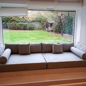 May include: A large, custom-built window seat featuring two rectangular gray cushions, two matching bolster pillows, and two square throw pillows. The seating area is integrated into a recessed alcove directly in front of a wide glass window.