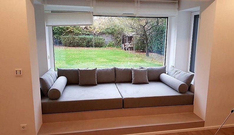 May include: A large, custom-built window seat featuring two rectangular gray cushions, two matching bolster pillows, and two square throw pillows. The seating area is integrated into a recessed alcove directly in front of a wide glass window.