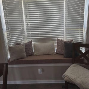 May include: A window seat cushion in a muted brown tone is topped with five decorative pillows in varying shades of cream, gray, and patterned fabric. The seat is positioned in front of a large bay window covered by white horizontal blinds.