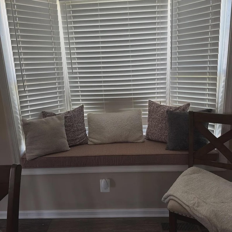 May include: A window seat cushion in a muted brown tone is topped with five decorative pillows in varying shades of cream, gray, and patterned fabric. The seat is positioned in front of a large bay window covered by white horizontal blinds.