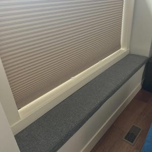 May include: A window seat cushion upholstered in a textured charcoal gray fabric, positioned within a white window frame. Above the cushion, a beige pleated cellular shade is partially lowered, filtering light through its horizontal honeycomb structure.