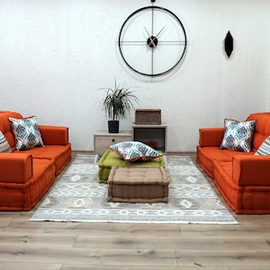 Puede incluir: Dos sof&aacute;s modulares naranjas con almohadas estampadas, un puf verde y un puf marr&oacute;n, y una alfombra con un patr&oacute;n geom&eacute;trico. Los sof&aacute;s est&aacute;n dispuestos uno frente al otro en un ambiente de sala de estar. Un reloj grande cuelga de la pared detr&aacute;s de los sof&aacute;s.