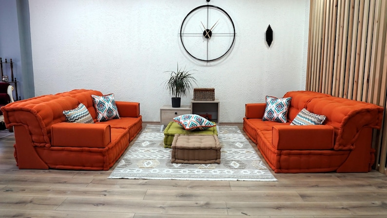 Puede incluir: Dos sof&aacute;s modulares naranjas con almohadas estampadas, un puf verde y un puf marr&oacute;n, y una alfombra con un patr&oacute;n geom&eacute;trico. Los sof&aacute;s est&aacute;n dispuestos uno frente al otro en un ambiente de sala de estar. Un reloj grande cuelga de la pared detr&aacute;s de los sof&aacute;s.
