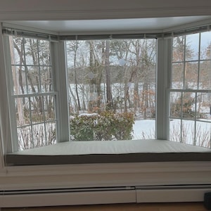May include: A white bay window featuring a built-in bench seat with a light gray cushion. The window panes offer a view of a snowy outdoor landscape with bare trees and a frozen body of water, framed by white wooden trim and horizontal blinds.