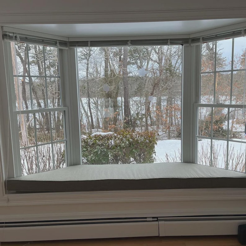 May include: A white bay window featuring a built-in bench seat with a light gray cushion. The window panes offer a view of a snowy outdoor landscape with bare trees and a frozen body of water, framed by white wooden trim and horizontal blinds.