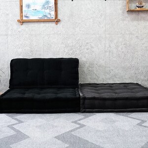 Puede incluir: Un sof&aacute; de coj&iacute;n de suelo de terciopelo de pana negro con un puf a juego. El sof&aacute; y el puf est&aacute;n en un sal&oacute;n con una alfombra gris, una planta en maceta y una estanter&iacute;a con elementos decorativos.