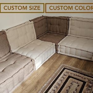 May include: A modular sectional sofa in shades of beige and brown. The square cushions are button-tufted. The text "CUSTOM SIZE" and "CUSTOM COLOR" are displayed above the sofa. A small rug rests on the wooden floor.