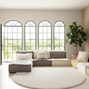 May include: A contemporary living room featuring modular sofas in grey and beige, with decorative cushions. A large, circular, white rug is on the floor. Tall arched windows and a potted plant enhance the room's design.