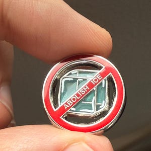 Puede incluir: Pin redondo plateado con borde rojo y la frase "ABOLISH ICE" en blanco. El pin presenta una imagen tachada de cubitos de hielo en el centro, con un tono azul claro.