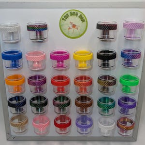 May include: A collection of small, clear plastic cylindrical containers with colourful, textured, screw-on lids arranged on a white magnetic board. A circular green logo with the text THE BUG DEN and an insect illustration is centred at the top.