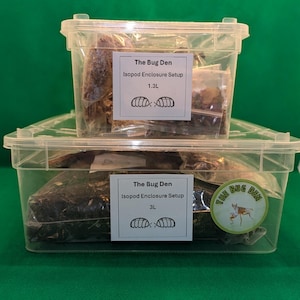 May include: Two clear plastic containers with lids, one stacked on top of the other. The top container is filled with brown and white materials, and the bottom container is filled with brown and green materials. Both containers have labels that say "The Bug Den Isopod Enclosure Setup" with a size in litres and a drawing of two isopods.