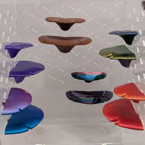 May include: A collection of ginkgo leaf-shaped objects in various colours including brown, purple, blue, green, red, and iridescent finishes. These items are arranged in a display, featuring smooth surfaces and distinct, fan-like shapes with central stems.
