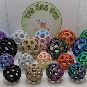 May include: A collection of 16 colourful, 3D printed, hollow, geometric balls. The balls are various shades of blue, green, purple, orange, pink, and brown. The balls are arranged in a circle on a white surface. The text "THE BUG DEN" is visible in the background.
