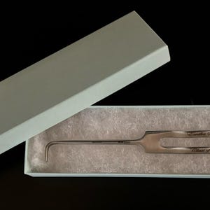 Könnte beinhalten: Ein silberfarbenes Lockpick-Werkzeug in einer hellblauen Geschenkbox mit passendem Deckel. Das Werkzeug hat ein gebogenes Ende und einen Griff mit Text. Die Box ist geöffnet und zeigt das Werkzeug auf einem Bett aus weißem Papier.