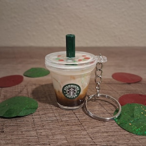 Könnte beinhalten: Ein Miniatur-Starbucks-Kaffeebecher-Schlüsselanhänger mit einem grünen Strohhalm und einem durchsichtigen Deckel. Der Becher hat einen silbernen Schlüsselanhängerring. Das Starbucks-Logo ist auf dem Becher sichtbar. Rote und grüne dekorative Formen umgeben den Becher.